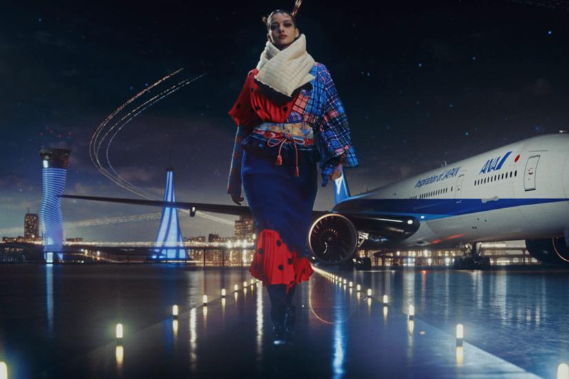 Fashion model wearing innovative Japanese streetwear walking on the runway next to an ANA aircraft at night.